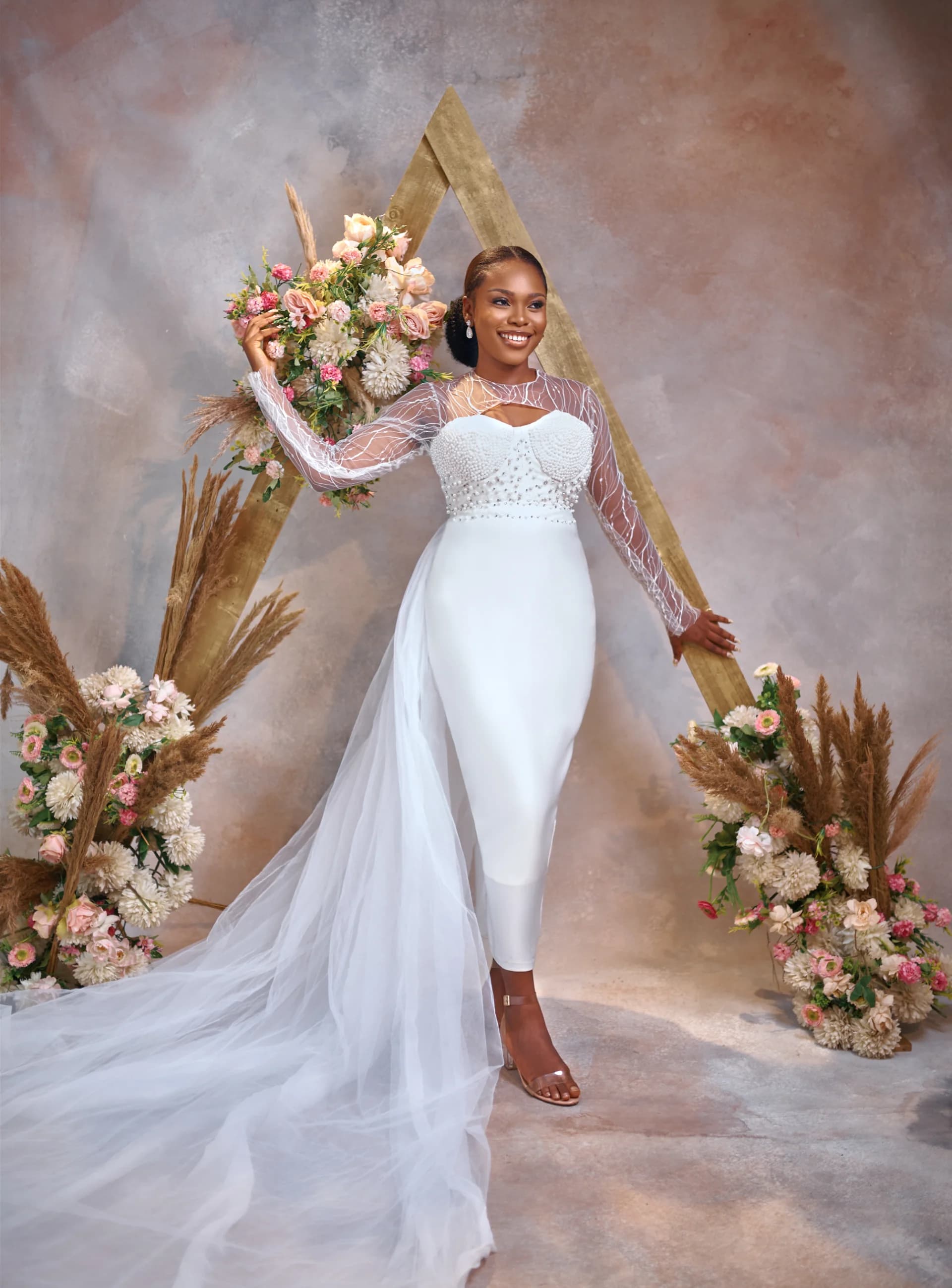 Bride holding a bouquet in a sheer-sleeve beaded gown, arch backdrop